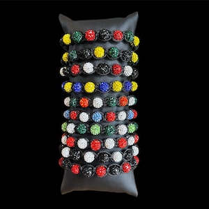 May include: A collection of beaded bracelets in various colors, including red, white, black, yellow, green, and blue. Each bracelet features a series of sparkling, round beads set in a black cord. The bracelets are displayed on a black stand against a black background.