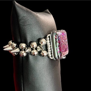 May include: A silver watch band with a pink and iridescent face and silver spikes. The band is on a black display stand.