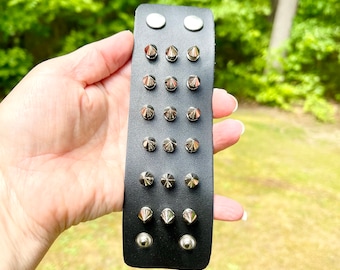 May include: A black leather bracelet with silver pyramid-shaped studs. The bracelet is wide and has multiple rows of studs.