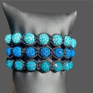 Puede incluir: Tres pulseras apiladas con cuentas de cristal azul en cordón negro. Las cuentas están graduadas en tamaño, con las cuentas más grandes en la parte inferior y las cuentas más pequeñas en la parte superior. Las pulseras son un degradado de azul, con la pulsera inferior siendo el azul más oscuro y la pulsera superior siendo el azul más claro.
