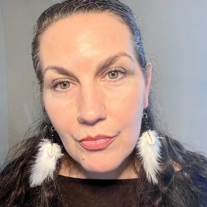 May include: A woman with long, dark hair is wearing a black top and white feather earrings with a shell accent. She has a nose ring.
