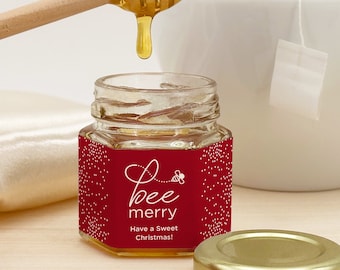 May include: A small glass jar of honey with a red label that says "bee merry" and "Have a Sweet Christmas!" The jar is on a white surface with a wooden honey dipper and a white teacup with a tea bag.