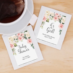 May include: Two white tea bag envelopes with floral designs. One envelope has the text "Baby Shower" and "For Jessica May 17, 2025". The other envelope has the text "It's a Girl!" and "Nicole's Baby Shower April 5, 2025".