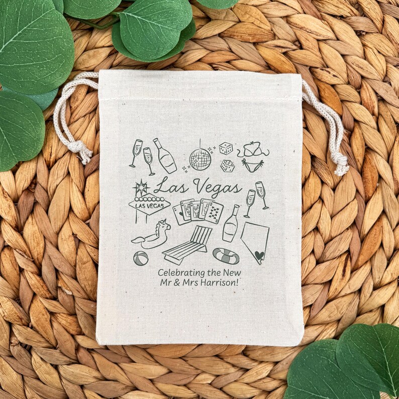 K&ouml;nnte beinhalten: Ein naturfarbener Canvas-Beutel mit Kordelzug und einem grauen Las Vegas-Design. Das Design umfasst Illustrationen von Sektgl&auml;sern, Flaschen, W&uuml;rfeln, Spielkarten und einem Schwimmreifen. Der Text lautet "Las Vegas" und "Celebrating the New Mr & Mrs Harrison!"