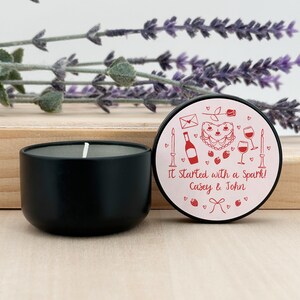 May include: A black candle tin with a pink lid. The lid has a red heart with a cake, wine bottle, and glasses. The text on the lid reads "It Started with a Spark! Casey & John".