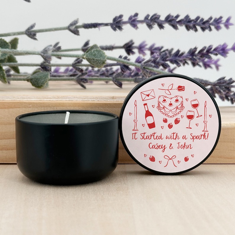 May include: A black candle tin with a pink lid. The lid has a red heart with a cake, wine bottle, and glasses. The text on the lid reads "It Started with a Spark! Casey & John".