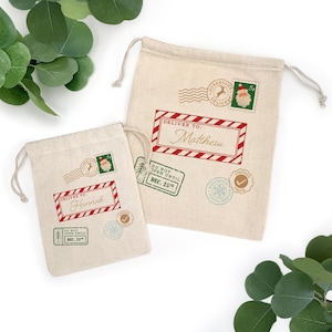 May include: Two beige drawstring gift bags with holiday-themed stamps and text. One bag reads "Hannah" and the other "Matthew" in a red and white frame. Other stamps include a Santa Claus stamp and a "Do Not Open Until Dec. 25th" stamp.