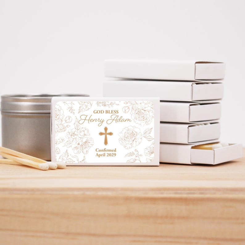 Puede incluir: Cajas de cerillas blancas con dise&ntilde;o floral y cruz. El texto en la caja de cerillas dice "GOD BLESS Henry Adam Confirmado Abril 2029". Una lata plateada y cerillas est&aacute;n al lado de las cajas de cerillas. Las cajas de cerillas est&aacute;n apiladas sobre una superficie de madera.