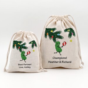 May include: Two beige drawstring bags with a pickle character playing pickleball under a Christmas tree design. One bag reads "Best Partner! Love, Ashley." The other says "Champions! Heather & Richard."