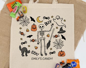 May include: Beige Halloween-themed tote bag with a skeleton, pumpkins, bats, spiders, and candy corn graphics. The text "EMILY'S CANDY!" is printed on the bag. Candy bars and treats are scattered around the bag.