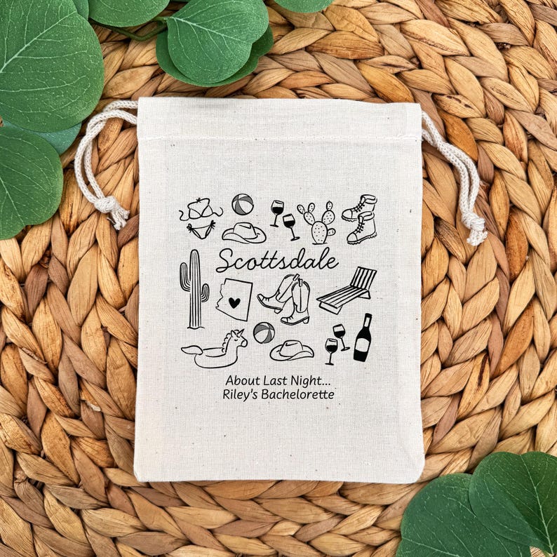 May include: A beige drawstring bag with a black illustration of Scottsdale-themed icons, including a cactus, cowboy boots, and wine glasses. The text "Scottsdale" and "About Last Night... Riley's Bachelorette" are also printed on the bag.