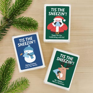 Peut inclure: Trois paquets de mouchoirs rectangulaires avec des graphismes de Noël. Un paquet bleu montre un bonhomme de neige avec le texte TIS THE SNEEZIN'! Happy Holidays Love, The Smiths. Deux paquets verts montrent le Père Noël et un renne.