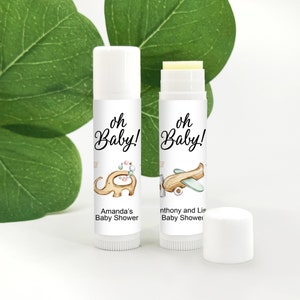 May include: Two white lip balm tubes with labels that read "oh Baby!" and baby shower details. One features an elephant illustration, the other an airplane. A white cap sits nearby. The background includes green leaves.
