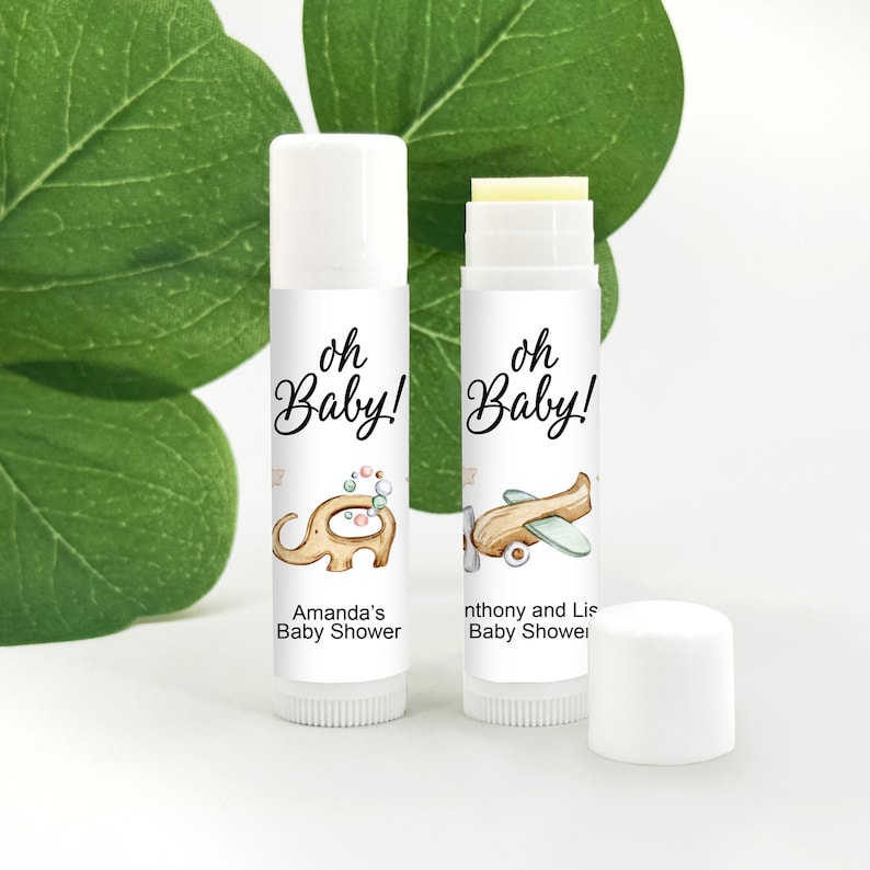 May include: Two white lip balm tubes with labels that read "oh Baby!" and baby shower details. One features an elephant illustration, the other an airplane. A white cap sits nearby. The background includes green leaves.