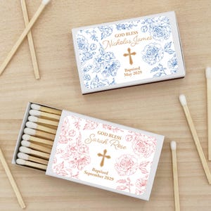 Puede incluir: Dos cajas de cerillas con dise&ntilde;os florales y texto personalizado. Una caja presenta patrones florales azules y el texto "God Bless Nicholas James", una cruz y "Bautizado mayo de 2029". La otra caja tiene patrones florales rosas y dice "God Bless Sarah Rose", una cruz y "Bautizado septiembre de 2029".