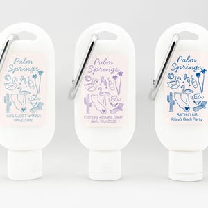 May include: Three white plastic bottles with a carabiner clip on top. Each bottle has a different design with the text "Palm Springs" and a different phrase below. The first bottle says "Girls Just Wanna Have Sun!" The second bottle says "Flocking Around Town! Girls Trip 2028". The third bottle says "Bach Club Riley's Bach Party".