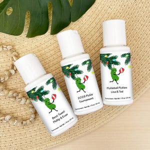 May include: Three white sunscreen bottles with white caps are arranged on a woven surface. Each bottle features a cartoon pickle playing pickleball under a Christmas tree design. Text includes "Best Team! Phillip & Evan", "2028 Pickle Tournament", and "Pickleball Pickles Lisa & Ted".