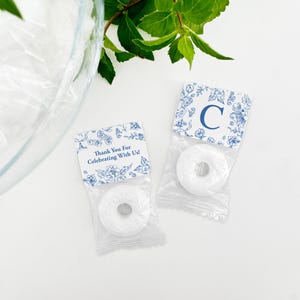 以下が含まれることがあります： 青い花柄のデザインと「Thank You for Celebrating With Us!」と「C」の文字が書かれた小さな白い袋が2つ。それぞれの袋には白い丸いミントキャンディーが入っています。