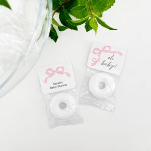 May include: Two white favor bags with pink ribbon designs. One bag has a tag that says "Sarah's Baby Shower" and the other says "oh baby!" Each bag contains a white round candy.