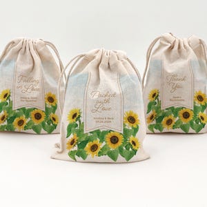 May include: Three beige drawstring bags with floral sunflower designs. Each bag has a different message: "Falling in Love", "Packed with Love", and "Thank You". The bags are decorated with yellow sunflowers and green leaves.
