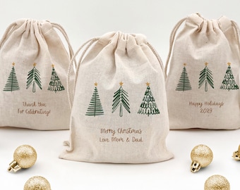 May include: Beige drawstring gift bags with green Christmas tree designs and gold star accents. Text on the bags reads "Thank You For Celebrating!", "Merry Christmas Love Mom & Dad", and "Happy Holidays 2029". Gold Christmas ornaments are scattered around the bags.