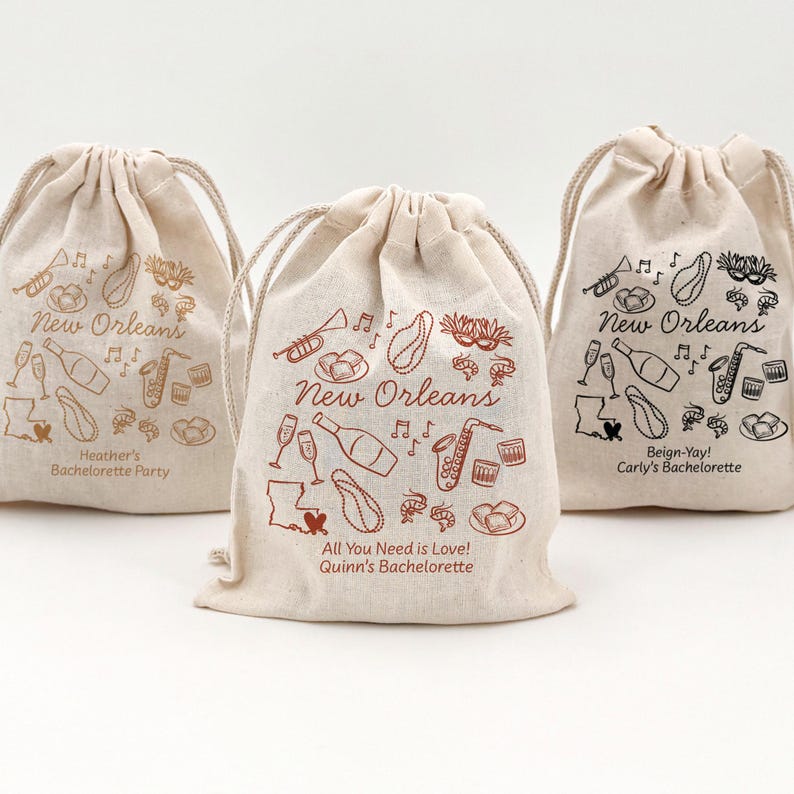 Three white drawstring bags (5" x 7" and 8" x 10") with black, gold, and terracotta New Orleans themed illustrations. The bags offer two lines of custom text and have a variety of New Orleans themed illustrations including beads, bourbon, music, and mardi gras mask.