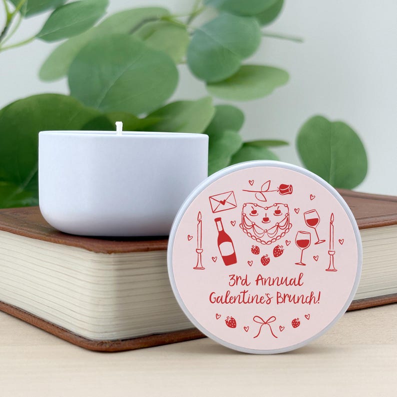 May include: White candle with a pink and red label that says "3rd Annual Galentines Brunch!" with illustrations of a heart-shaped cake, wine glasses, a bottle of wine, a rose, and candles.
