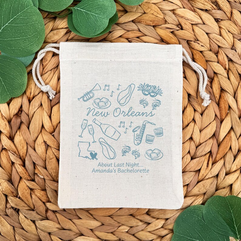 May include: A beige drawstring bag with a light blue illustration of New Orleans icons, including a trumpet, musical notes, and a saxophone. The text "New Orleans" and "About Last Night... Amanda's Bachelorette" are also printed on the bag.