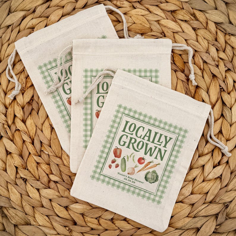 May include: Three off-white drawstring bags with a green gingham border and the words "Locally Grown" printed on them. The bags feature illustrations of vegetables and the text "Violet's Baby Shower 06.06.23".