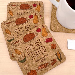 May include: Cork coasters with a Thanksgiving theme. The coasters feature illustrations of a roasted turkey, apples, pears, and other festive foods. Text on the coasters reads "Let's Get Stuffed" and "Thanksgiving 2029".