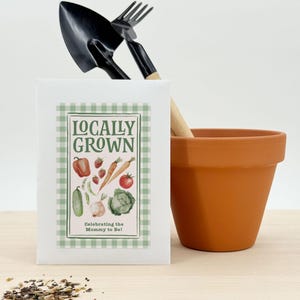 Puede incluir: Un sobre de semillas blanco con un borde de cuadros verdes y las palabras "Locally Grown" e ilustraciones de verduras. Una pequeña paleta y un tenedor de jardinería están detrás del sobre, con una maceta de terracota a la derecha. El texto "Celebrating the Mommy to Be!" está en la parte inferior.