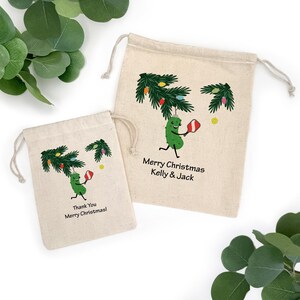 May include: Two beige drawstring bags with Christmas-themed illustrations. One bag features a green pickle character holding a red and white ornament under a decorated tree. The text "Merry Christmas Kelly & Jack" is printed on the bag. The smaller bag says "Thank You Merry Christmas!"
