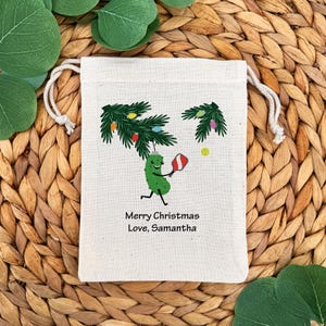 May include: A cream-colored drawstring gift bag featuring a cartoon pickle playing pickleball under a Christmas tree. The bag has the text "Merry Christmas Love, Samantha". The background is a woven texture with green leaves.
