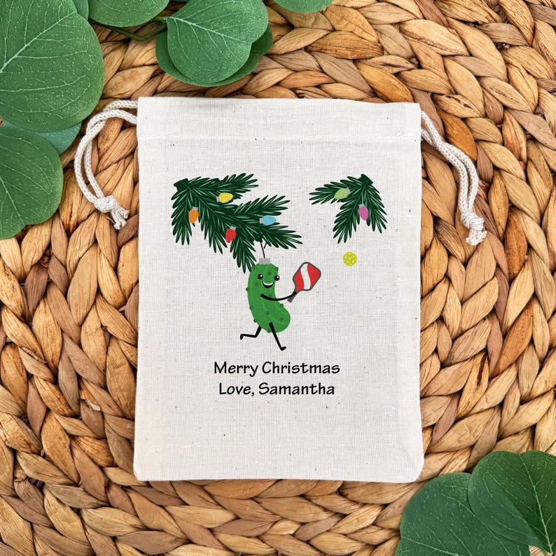 May include: A cream-colored drawstring gift bag featuring a cartoon pickle playing pickleball under a Christmas tree. The bag has the text "Merry Christmas Love, Samantha". The background is a woven texture with green leaves.