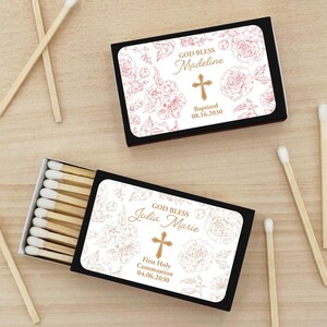 Puede incluir: Dos cajas de cerillas negras con dise&ntilde;os florales blancos. Una caja dice "GOD BLESS Madeline" con una cruz y "Baptized 08.16.2030". La otra dice "GOD BLESS Julia Marie" con una cruz y "First Holy Communion 04.06.2030."