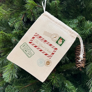 May include: A beige drawstring gift bag with red and green postage-themed graphics. The bag reads "Deliver To: The Good Child" and includes a stamp with Santa Claus. The bag is decorated with other postage-themed stamps.