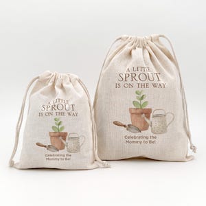 May include: Two beige drawstring bags with the text "A Little Sprout Is On The Way" and a watercolor illustration of a plant in a pot, a watering can, and a trowel. The bags are for celebrating the Mommy to Be!