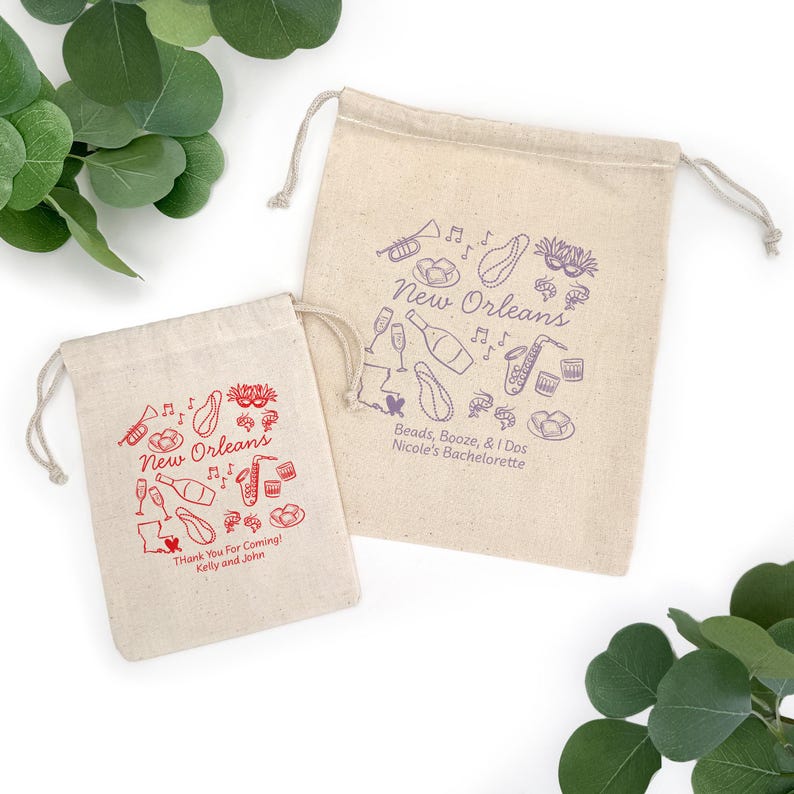 May include: Two beige drawstring bags with New Orleans-themed illustrations. One bag has red graphics and text, including "Thank You For Coming!" The other has purple graphics and text, including "Beads, Booze, & i Dos".