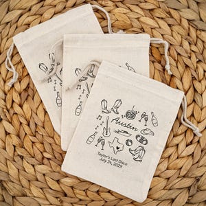 May include: Three beige drawstring bags with black illustrations of cowboy boots, musical notes, and bottles. One bag has the text "Austin" and "Taylor's Last Disco July 24, 2029". The bags are on a woven surface.