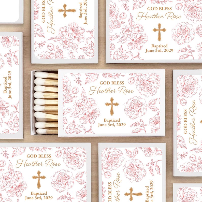 White matchboxes labeled with a floral design and text that reads "GOD BLESS", custom text for the child's name, baptized, and date. The boxes feature a cross and are surrounded by a floral pattern. The matches are visible in an open box.