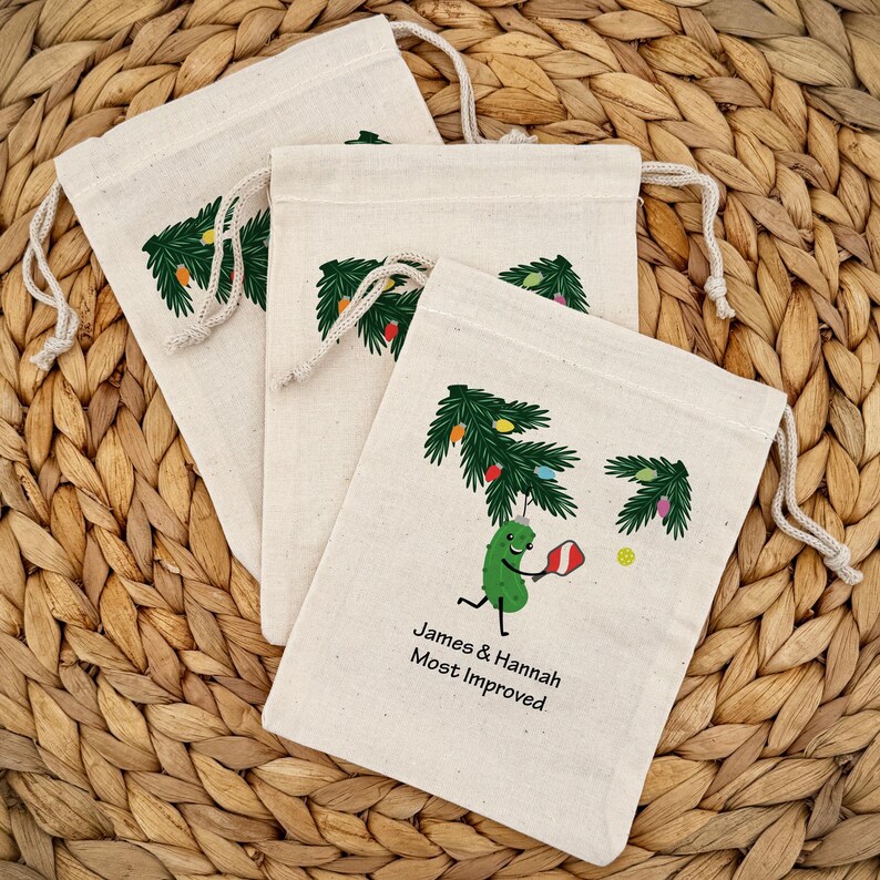 May include: Three off-white drawstring bags with a green Christmas tree design and colorful lights. One bag features a cartoon pickle playing pickleball with the text "James & Hannah Most Improved". The bags are on a woven surface.