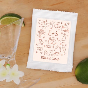 Puede incluir: Una bolsa de regalo blanca con una etiqueta rosa que presenta un diseño de temática nupcial con ilustraciones de corazones, anillos, copas de champán y una cámara. La etiqueta también incluye el texto "E+S" y "Ethan & Sarah".