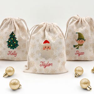 May include: Three beige drawstring gift bags with Christmas-themed designs. One bag features a Christmas tree, another Santa Claus, and the third an elf. Each bag has a name in red script: Kelly, Taylor, and Ryan. Gold Christmas ornaments are scattered around.