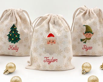 May include: Three beige drawstring gift bags with Christmas-themed designs. One bag features a Christmas tree, another Santa Claus, and the third an elf. Each bag has a name in red script: Kelly, Taylor, and Ryan. Gold Christmas ornaments are scattered around.