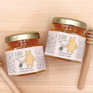 May include: Two small glass jars filled with honey, decorated with Winnie the Pooh and Piglet characters. The labels read "A Little Honey is on the Way" and "Celebrating the Parents to BEE!" One jar also has the text "Baby Peterson Coming this June!"