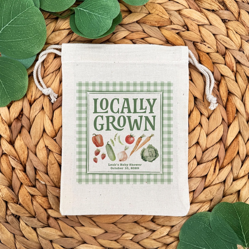 May include: A cream-colored drawstring bag with a green gingham design and the words "Locally Grown" printed on the front. The design includes illustrations of vegetables. The text "Leah's Baby Shower October 10, 2020" is printed below the vegetables.