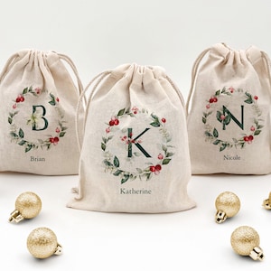 May include: Three beige drawstring gift bags with initial letters inside a wreath of green leaves and red berries. The names "Brian", "Katherine", and "Nicole" are printed below the bags. Gold glitter ornaments are scattered in front of the bags.