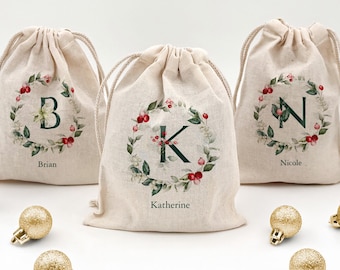May include: Three beige drawstring gift bags with initial letters inside a wreath of green leaves and red berries. The names "Brian", "Katherine", and "Nicole" are printed below the bags. Gold glitter ornaments are scattered in front of the bags.