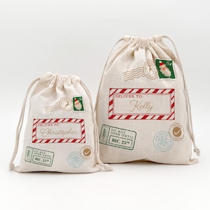 May include: Two beige drawstring gift bags with red and green postage-themed designs. One bag says "Deliver to: Christopher" and the other says "Deliver to: Kelly". Both bags have "Do Not Open Until Dec. 25th" printed on them.