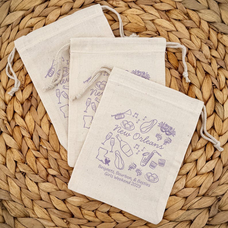 May include: Three beige drawstring bags with purple illustrations of New Orleans icons, including a saxophone, champagne glasses, and beignets. The text "New Orleans" and "Beignets, Bourbon, & Besties Girls Weekend 2029" are also printed on the bags.